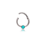 Introducing the "Turia" by Kaya Movement: a minimalist silver-finish septum jewelry piece crafted from 316L stainless steel. This refined and elegant hoop nose ring features a textured design with a small turquoise gemstone at its center, perfect for the nose, tragus, or lobe. It has a sleek design measuring 1.2mm and includes an open, adjustable end for easy insertion and removal. The vibrant blue accent of the gemstone stands out beautifully against its polished silver finish.