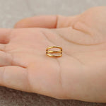 A close-up image of a person holding the Minimalist Gold PVD 316L Steel Septum "Trion" by Kaya Movement, designed with a sleek triple ring effect and subtle elegance. The hand is centered while the shiny surface of the 1.2mm, 8mm diameter ring reflects light against an out-of-focus greyish textured background.