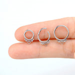 A hand holds three Zuri Minimalist Mandala-Inspired Septums from Kaya Movement, designed in 316L steel with a silver finish, against a plain background. Each piece features an 8mm diameter and minimalist design with a small heart at the bottom, accented by two dot decorations. One septum piece is slightly open to showcase the clasp mechanism.