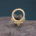 A minimalist, intricate septum ring from Kaya Movement's "Zura" collection, finished in 316L PVD Gold Steel, lies against a rustic, textured background. The ring features delicate scrollwork detailing with a central heart design at the bottom, accented by tiny gold beads on either side. Available in diameters of 6mm, 8mm, and 10mm with a gauge of 1.2mm, it beautifully embodies the elegance of mandala-inspired jewelry.