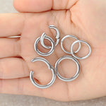 A person holding six Kaya Movement Aurel Simple Ear Rings in their hand, each crafted from 316L steel with a gleaming silver finish. The ear rings come in varying sizes of 2mm and 3mm gauges, with diameters of 8mm, 10mm, 12mm, and 14mm. Arranged loosely on the palm over a tan-colored surface, these rings showcase the refined allure of minimalist elegance.