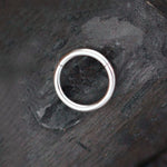 A single Aurel Simple Ear Ring in 316L Steel Silver Finish from Kaya Movement rests on a textured, dark gray surface. This earring, available in gauges of 2 or 3mm and diameters of 8, 10, 12, or 14mm, embodies minimalist elegance with its slightly open design revealing the clasp mechanism.