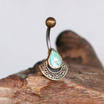 Presenting the Lumi Navel Jewelry by Kaya Movement, a boho-style ethnic piece featuring a curved barbell and round top ball. This unique handcrafted festival gem boasts a pear-shaped abalone shell set in an ornately detailed bronze setting, showcasing the iridescent blue and green hues of the abalone shell on wood.