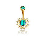 The Solis Navel Piercing by Kaya Movement showcases a geometric golden design with boho shine. The exquisite piece features a small, round green-blue abalone shell at the top and a larger matching gemstone at the bottom, encircled by a decorative array of small gold beads. Its intricate craftsmanship and gleaming gold finish make this striking accessory stand out beautifully against a white background.