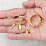 A hand holding several Zuri Tunnels by Kaya Movement, gold-colored ear plugs and tapers for stretching, crafted from 316L surgical steel with a Gold PVD finish. The items vary in size, featuring shiny metallic surfaces, and are displayed against the palm on a light textured background. Perfect minimalist jewelry for any enthusiast. Available in 6mm and 16mm sizes.