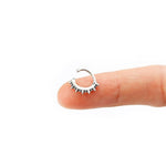 A person's finger holds a small, Silver "Stera" Septum: Star's Glow earring by Kaya Movement with a spiked design. The plain white background emphasizes the earring's detail and relative size, making it ideal for stylish nose, helix, tragus, lobe, or conch piercings.