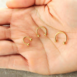 A close-up of a hand holding three small Kaya Movement 316L Steel PVD Gold Circular Barbell Septum "LUMA" piercings, with diameters of 6mm, 8mm, and 10mm. These minimalist pieces feature a ball on each end and are artfully displayed in the open palm. The slightly blurred background enhances the refined elegance of these nose, tragus, and lobe accessories.