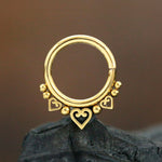 A close-up image showcases a Kaya Movement 316L Steel PVD Gold Mandala Design Septum "Natura" nose ring, featuring intricate designs. The mandala is adorned with three small heart-shaped embellishments and tiny gold bead accents. The PVD gold finish and blurred background enhance the focus on this detailed jewelry piece, highlighting its nature and spirituality-inspired design.