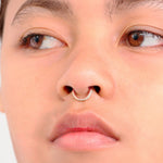 Close-up of a person's face showcasing a Kaya Movement Hinged Septum Ring Clicker 16g Cubic Zirconia with a gold ring adorned with small, clear stones. The person's eyes are looking to the side, their expression neutral. The background is plain and white.