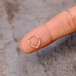 A close-up image of a human finger holding a small, heart-shaped gold body jewel. The delicate and minimalistic Hinged Septum Ring by Kaya Movement contrasts with the textured, slightly blurred background.