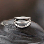 A gold PVD 316L steel piece from Kaya Movement, known as the "Trion" with a triple ring effect, rests on a dark, textured surface. This minimalist septum accessory features three sleek and slightly curved bands connected at the back for an elegant and modern look. The refined design boasts a 1.2mm thickness and 8mm diameter, exuding subtle elegance for nose wear.