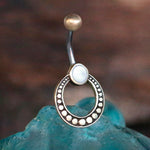 The Azmi Navel Jewelry by Kaya Movement, featuring a Boho Ethnic design with a mother of pearl-like white gem set in a circular, dotted brass outer ring, stands upright against a blurred, earthy background. The piece includes a curved surgical steel barbell with a ball at the end, which is partially visible behind a blue object.