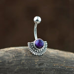The Nala Navel Jewelry by Kaya Movement, a boho ethnic piece made of 316L surgical steel, features an Inca-inspired half-moon design with a central purple amethyst. Positioned against a dark background, the jewelry rests on a textured wooden surface.