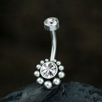 A close-up shot features Kaya Movement's Zirka Navel Jewelry - Boho Essence with Sparkling Zircon, crafted from silver 316L surgical steel. The exquisite belly button ring showcases two clear gemstones; a smaller one sits at the top, and the larger stone is nestled in a decorative flower-like design at the bottom. This elegant piece of natural and bohemian navel jewelry is displayed on a dark, textured surface.
