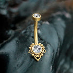 Close-up of the Zarina Navel Piercing by Kaya Movement, showcasing its bohemian elegance with a golden mandala floral design. The intricate craftsmanship highlights a central sparkling zircon, surrounded by five small gold beads and three heart-shaped accents, all gracefully positioned on a dark, smooth surface.
