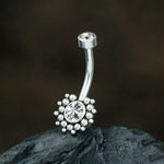 Close-up image of the Sira Navel Piercing by Kaya Movement, featuring a modern silver finish with two sparkling zircon gemstones. One gemstone is set at the top of the straight bar, while a larger, flower-like design with a central gemstone surrounded by silver beads adorns the bottom. This exquisite piece, designed to be worn on a pierced belly button (10mm - 1.6mm gauge 14g), is displayed on a dark, textured surface.