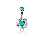 The Kazi Navel Piercing by Kaya Movement, featuring a geometric silver design with a bohemian reflection, showcases the vibrant shades of blue, green, and hints of purple in its abalone shell inlay. This stainless steel belly button ring includes a small abalone gem at the top and a larger one centrally set within an intricate circular beaded pattern, making it perfect for enhancing any boho outfit. This stylish navel piercing is available in gauge 1.6mm (14g) with a length of 10mm.