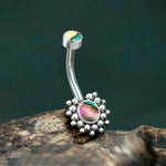 The Kazi Navel Piercing from Kaya Movement, featuring a sleek geometric silver design, showcases a small top ball with an opalescent finish and a larger bottom charm inlaid with abalone shell surrounded by silver beads. Perfectly set on a rough, natural wooden surface, this stainless steel piercing (gauge 1.6mm 14g - 10mm) is an ideal accessory for boho outfits that celebrate eclectic and natural style.