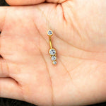 A close-up image captures a human hand holding the Kaya Movement Xane 1.6mm Navel Piercing, boasting a brilliant gold finish. The surgical steel piece is adorned with four sparkling zircons arranged vertically along its curve. The palm of the hand elegantly serves as the background for this luxurious navel piercing jewelry.