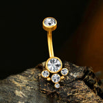 The Lysor 1.6mm Navel Piercing by Kaya Movement features a gold curved barbell adorned with six bright zircons of varying sizes arranged in a chic mandala pattern. This elegant and sparkly design, showcased on a textured rock background with a dark gradient, is perfect for adding a touch of luxury to both boho outfits and navel piercings alike.
