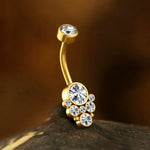 A close-up image showcases the Kaya Movement's Octra 1.6mm Navel Piercing - Mandala Chic Brilliance, featuring a curved barbell design in a gold finish. Adorned with eight stunning white zircon gemstones of varying sizes, including a larger central one surrounded by smaller gems, the piercing exudes sublime elegance. The background is blurred and dark.