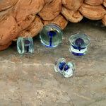 A close-up image of four small Kaya Movement's Psylia Plugs with blue mushroom glass designs, featuring clear and blue swirls, lying on a gray marble surface. In the background, there is a wooden textured object reminiscent of a psychedelic festival accessory. Available in sizes 6mm to 16mm (2g to 5/8"), these plugs encapsulate a true psychedelic festival vibe.