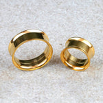 Two gold plugs from Kaya Movement's Zuri Tunnel collection, meticulously crafted from 316L surgical steel with a Gold PVD finish, rest on a light gray, textured surface. The plug on the left is larger and slightly thicker at 16mm compared to the smaller 6mm one on the right. Both plugs exemplify minimalist design with their smooth, shiny finish.