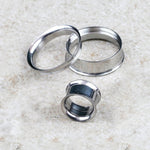 Three Kaya Movement Zura Tunnel ear plugs of varying sizes are scattered on a textured white surface. One ear plug stands upright while the other two lie on their sides, showcasing the minimalist charm and unique angles of these 316L surgical steel jewelry pieces in silver finish.