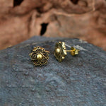 Close-up of a pair of intricate, Zenaya Brass Ear Studs by Kaya Movement. Featuring a floral design reminiscent of mandala lotus patterns, these earrings have a central bead with petal-like details extending outward. They are displayed on a textured rock surface with a blurred background.