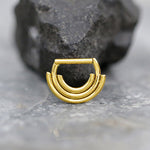 A close-up photo of an elegant Anneau de Septum Or 8mm Triple Empilé by Kaya Movement, crafted from Premium Surgical Steel and resting on a marble surface. The hypoallergenic and lightweight ring showcases a three-layered arc design, with a large piece of dark stone in the background, exuding contemporary sophistication.