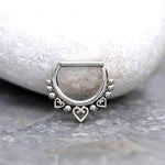 A silver Piercings Nez 8mm Septum Clicker Argent by Kaya Movement, meticulously designed with a row of small silver beads and three heart-shaped accents at the bottom. The elegant piercing is displayed on a smooth white stone, set against a gray marbled surface.