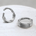 A close-up image of two Clicker pour Septum 8mm en Acier Chirurgical 16g - Anneau Épuré Triple Lignes by Kaya Movement, resting on a white surface. One piece is standing upright, displaying its open clasp, while the other lies flat. The jewelry features a minimalist and polished design with a series of parallel grooves along the band.