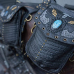 Close-up of the SHIPIBO Tribal Apocalyptic Leather Belt & Shoulder Bag by Kaya Movement, showcasing its black leather adorned with brass rivets and intricate designs. Reminiscent of Wasteland hip bag styles, the flap features a blue labradorite stone centerpiece surrounded by grey textured panels and gold-colored metallic accents, including a decorative cross at the top.