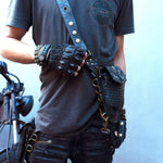 A person dressed in a grey T-shirt and black leather accessories, including studded gloves, multiple belts, straps, and a Kaya Movement SHIPIBO Tribal Apocalyptic Leather Belt & Shoulder Bag featuring Labradorite, stands in front of a motorcycle. The detailed leather gear showcases studs, rivets, and multiple compartments.
