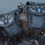 A close-up view of two intricately designed SHIPIBO Tribal Apocalyptic Leather Belt & Shoulder Bags by Kaya Movement, featuring metal clasps and Labradorite gemstone decorations. The bags are strapped onto a piece of weathered, rustic metal equipment with brass rings and fastenings, evoking the rugged charm suited for dystopian festivals. The background is a neutral grey wall.