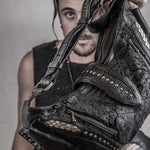 A person with long hair and a beard partially visible in the background holds up the rugged and stylish Nebula Unisex Leather Off Shoulder Cross Body HandBag from Kaya Movement, featuring intricate detailing, studs, and zippers that showcase its design. The plain and neutral backdrop contrasts sharply with the dystopian festival vibe of this accessory.