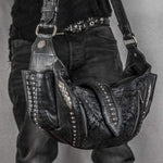 A person dressed in dark clothing holds a Nebula Unisex Leather Off Shoulder Cross Body HandBag by Kaya Movement. This custom black leather bag boasts intricate details, including metallic studs, textured patterns, and side panels akin to reptile skin. The individual's belt also features metal studs, perfectly complementing the edgy design of the handbag, making it ideal for dystopian festivals.