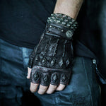 A person sporting a black, fingerless, textured glove adorned with metal studs and intricate designs rests their hand on their hip. They pair it with dark pants and an edgy KRAKO Reptilian Spine Cuff Bangle in Black from Kaya Movement, showcasing an adjustable studded bracelet embellished with multiple spikes. The overall aesthetic is edgy and punk-inspired.