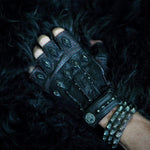 A close-up image of a hand wearing a black fingerless leather glove with metallic studs and red stitching. The wrist is adorned with multiple silver spiked bracelets and a Kaya Movement KRAKO Reptilian Spine Cuff Bangle in Black, adjustable for unisex wear, against a background featuring a black, furry texture.