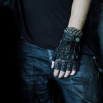A person is wearing black fingerless gloves adorned with studs and dark beads, a spiked bracelet, a black T-shirt, and distressed black leather pants with zippers. They are also sporting the KRAKO Reptilian Spine Cuff Bangle by Kaya Movement, made using the lost wax process. They are standing against a dark background with their arm resting by their side.