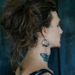 A person with long, loosely tied hair is shown in a side profile. They are wearing Kaya Movement's PROMETHEUS Medium Biomechanical Hoop Earrings made of 925 silver (18 gauge) and have a nose ring. A butterfly tattoo is visible on their neck. The background is dark and blurred.