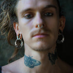 A person with light skin and long wavy hair is wearing Kaya Movement's PROMETHEUS Medium Biomechanical Hoop Earrings in 925 Silver, 18 gauge, along with a septum ring and displaying tattoos on their neck, which include a butterfly and a moth. The background is dark and out of focus, bringing attention to the subject's face and jewelry.