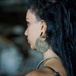 A person with long, dark hair is shown in profile. They are wearing a large, intricately designed gold boho statement earring from Kaya Movement's SAYAGATA Oversized Tribal Hoop Disc Earrings collection in 16 gauge, along with a smaller hoop earring and a nose ring. The background is blurred.