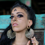 A person with long dark hair poses, resting their head against a teal wooden backdrop. They have intricate black facial markings and wear two large CIRCUIT Oversized Visionary Disc Hoop Earrings in Gold by Kaya Movement. They also have a nose ring and wear dark eye makeup, highlighting their eyes.