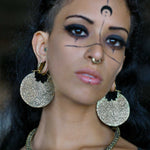 A person with PANCHA Oversized Tribal Shipibo Disc Earrings in Gold by Kaya Movement, along with a septum ring and a lip ring, has black face paint designs extending from their nose to cheeks, forehead, and chin. They have dramatic eye makeup and a pensive expression.