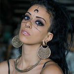 A person with elaborate facial markings, including a small crescent moon on their forehead, stares intently at the camera. They have dark eye makeup, a nose ring, a chunky necklace, and wear a black strap top. They are also wearing the AREVALO Shipibo Oversized Disc Hoop Earrings in Gold by Kaya Movement.