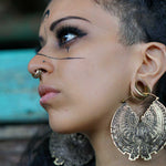 Close-up side profile of a person with intricate black facial markings, wearing the Kaya Movement DARMA Biomechanical Clicker Hoop Septum Nose Ring in Gold (16 gauge) and ornate large round earrings. The person's makeup is bold, featuring winged eyeliner, and the background is a blurred blend of turquoise and neutral tones.