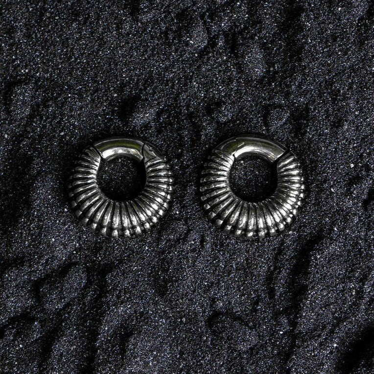 A pair of TRILOBITE Small Biomechanical Clicker Ear Weights in Silver Brass (2 gauge) from Kaya Movement are placed on a textured black surface. The dark background accentuates the shiny, reflective nature of these biomechanical earrings.