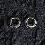 A pair of TRILOBITE Small Biomechanical Clicker Ear Weights in Silver Brass (2 gauge) from Kaya Movement are placed on a textured black surface. The dark background accentuates the shiny, reflective nature of these biomechanical earrings.