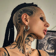 A person with intricate tattoos poses while wearing large circular metallic earrings, a septum ring, and black nail polish. Their dramatic eye makeup and red lighting create a bold and intense atmosphere. They touch their lips with a confident gaze.